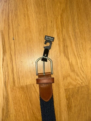 Marinblått flätat bälte med skinn - Snyggt marinblått flätat bälte med detaljer i brunt skinn och silverfärgat spänne. Perfekt accessoar för att lyfta din outfit. Längd 95 cm och bredd 34 mm. Klassisk design som passar till många olika stilar.