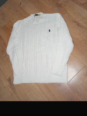Vit kabelstickad tröja Polo Ralph Lauren - Kabelstickad vit tröja från Polo Ralph Lauren med rund halsringning och den klassiska broderade logotypen på bröstet. Tröjan har långa ärmar och ribbade muddar vid ärmslut och nederkant. Perfekt för en clean och stilren look.