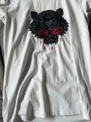 Vit Kenzo t-shirt med tigerbrodyr - Säljer en vit t-shirt från Kenzo med ikonisk svart och röd tigerbrodyr och texten 'KENZO PARIS' på bröstet. Klassisk passform och korta ärmar. Tillverkad i mjuk bomull som känns skön mot huden. Perfekt för dig som vill ha en statement-tee med lyxig känsla. (HÄMTAS I GBG)