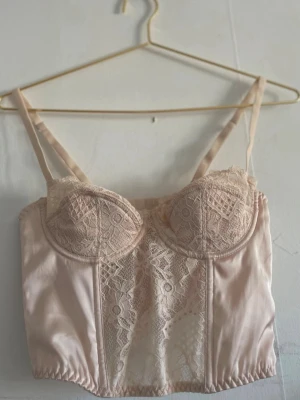 Ljusrosa spetskorsett/linne med satin Urban Outfitters - Säljer en ljusrosa linne med tunna axelband, vadderade kupor och vacker spetsdetalj framtill. Storlek S/M. Materialet är en kombination av satin och spets som ger en lyxig känsla. Perfekt att styla under en skjorta eller som statement-plagg.