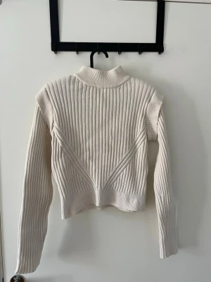 Tröja / top - Säljer en vit- beige ribbstickad tröja från NA-KD med hög krage och långa ärmar. Tröjan har ett snyggt mönster med diagonala ribbor framtill och är gjord i mjukt stickat material. Lätta vaddar i axlarna. Stl. xxs- xs