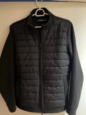 Svart pufferjacka från dressmann - Stilren svart pufferjacka från dressmann med quiltad framsida och släta ärmar. Jackan har dragkedja framtill, ståkrage och två sidofickor. Perfekt för sköna vårdagar och enkel att matcha med olika outfits.