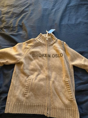 Beige ziptröja från Broken Oslo - Säljer en beige ziptröja från Broken Oslo med hög krage och ribbade muddar. Tröjan har två fickor framtill och texten 'BROKEN OSLO' tryckt över bröstet. Perfekt för dig som gillar streetwear och vill ha en stilren men avslappnad look.