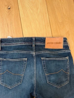 Blå jeans från Jacob Cohën - Snygga blå jeans från Jacob Cohën med klassisk femficksmodell och kontrastsömmar. Jeansen har en läderpatch med logga bak i midjan och raka ben. Materialet är denim i bomull med en mjuk känsla. Perfekta för en avslappnad och trendig stil.