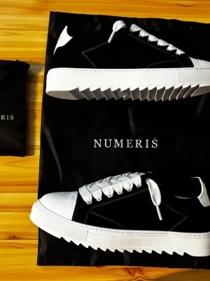 Svarta och vita sneakers från Numeris - Snygga sneakers från Numeris i svart mocka med vita detaljer och grov, räfflad sula. Skorna har vita skosnören, vit tå och häl samt kontrastsömmar. Perfekta för dig som vill sticka ut med en modern och trendig look.