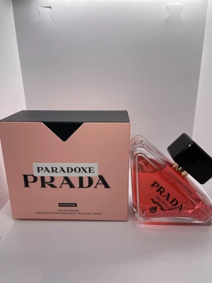 Prada Paradoxe Intense EdP - Prada Paradoxe Intense Eau de Parfum har en unik trekantig glasflaska med svart lock och rosa-orange doftvätska. Förpackningen är ljusrosa med svarta detaljer och tydlig Prada-logga. Parfymen är påfyllningsbar och har en modern, lyxig design som sticker ut på hyllan.
