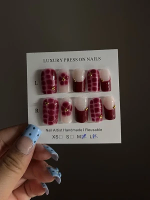Vinröda press-on naglar med mönster - Lyxiga press-on naglar i vinrött och nude med handmålat krokodil- och blom-mönster samt gulddetaljer. Naglarna har en rak, kort form och är tillverkade i plast. Perfekt för dig som vill ha en unik och trendig look.