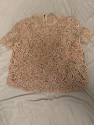 Beige spetsblus med dragkedja - Säljer en beige blus i spets med korta ärmar och snygg dragkedja i ryggen. Blusen har ett detaljerat spetsmönster som ger en romantisk vibe och är perfekt för att lyfta vilken outfit som helst.