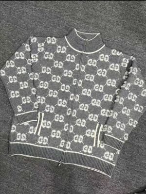 Grå stickad ziptröja från Gucci - Snygg grå stickad ziptröja från Gucci med vit GG-logga över hela plagget. Tröjan har ribbade muddar, hög krage och vita detaljer vid dragkedja, ärmslut och nederkant. Perfekt för dig som gillar exklusiv streetstyle och vill sticka ut.