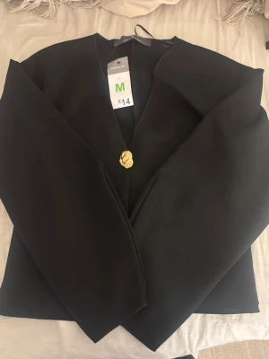 Svart kavaj med guldig knapp Primark - Stilren svart kavaj från Primark i storlek M. Kavajen har en rak passform och stängs med en dekorativ guldig knapp framtill. Materialet är slätt och ger ett elegant intryck. Perfekt för dig som vill ha en enkel men snygg look.