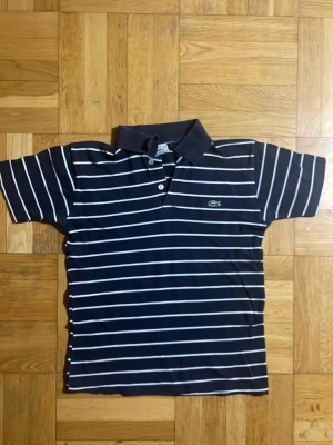 Marinblå randig piké från Lacoste - Snygg marinblå pikétröja från Lacoste med vita horisontella ränder och klassisk krage. Tröjan har korta ärmar, två knappar framtill och den ikoniska krokodilloggan broderad på bröstet. Tillverkad i mjuk bomull för en bekväm känsla.