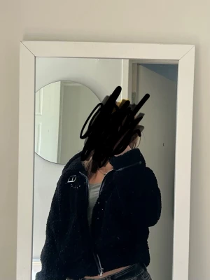 Svart teddyjacka med dragkedja - Mysig svart teddyjacka med hög krage och dragkedja framtill. Jackan har en boxig passform och en dekorativ spänndetalj vid kragen. Perfekt för att hålla värmen under kyliga dagar.