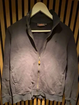 Mörkblå ziptröja från Morris - Mörkblå ziptröja från Morris med hög krage och diskret logga på bröstet. Tröjan har två fickor framtill, ribbade muddar och dragkedja hela vägen. Perfekt för en avslappnad och stilren look.