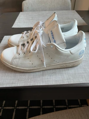 Adidas Stan Smith vita sneakers storlek 42 - Klassiska vita Adidas Stan Smith sneakers med blå detaljer och perforerade ränder på sidorna. Skorna har lågt skaft, vita snören och Adidas-logga samt Stan Smith-tryck på hälen. Tillverkade i läder med gummisula. Perfekta för en clean och tidlös stil.
