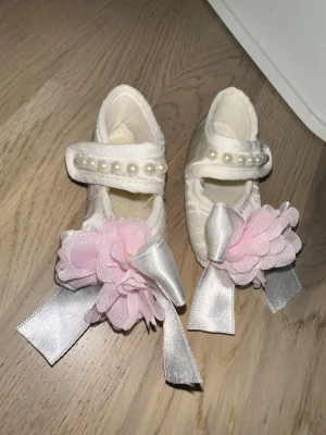 Vita babyskor med pärlor och rosett - Söta vita babyskor med dekorativa pärlor på remmen och stora rosa tygblommor framtill. Skorna har även silvriga band och är tillverkade i tygmaterial. Perfekta för speciella tillfällen eller dop.