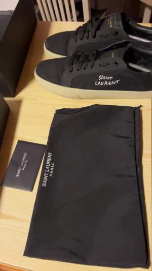Svarta Saint Laurent sneakers - Svarta sneakers från Saint Laurent med vit logga på sidan och beige platt sula. Skorna har snörning och rund tå, tillverkade i canvas och skinn. Medföljer originalkartong och dustbag med Saint Laurent Paris-tryck. Perfekt för dig som gillar stilrena och lyxiga sneakers. 