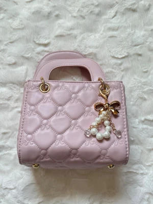 Ljusrosa quiltad handväska med pärldetalj - Supersöt ljusrosa handväska i quiltat mönster med guldfärgade metalldetaljer och ett dekorativt pärlhänge med rosett. Väskan har två handtag och dragkedja på insidan. Perfekt för dig som gillar en feminin och chic stil.