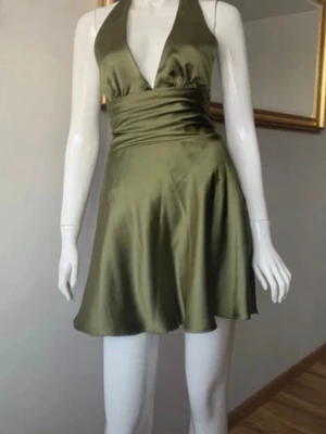 Olivgrön omlottklänning i satin - Snygg olivgrön kort klänning i satin med omlottmodell och djup v-ringning. Klänningen har öppen rygg och knyts med en stor rosett baktill. Perfekt för dig som vill ha en elegant och trendig look.