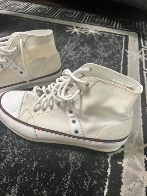 Vita höga sneakers med röd detalj - Snygga vita höga skor från Gucci i canvas med vita snören. Skorna har en tunn rand och klassisk gummisula. Perfekta för en clean och stilren look. 