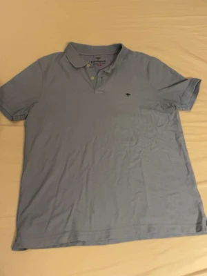 Ljusblå pikétröja  - Säljer en klassisk ljusblå pikétröja från Fynsh hutton med liten broderad logga på bröstet. Tröjan har korta ärmar, krage och knappar framtill. Perfekt för en avslappnad och stilren look.