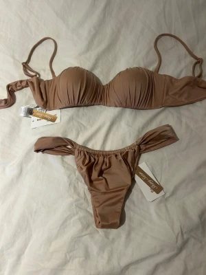 Beige bikini från Calzedonia - Snygg beige bikini från Calzedonia med vadderad bygel-bh och matchande trosa med knyt i sidorna. Båda delarna har en lätt glansig finish och enkel, stilren design. Perfekt för strandhäng eller pooldagar.