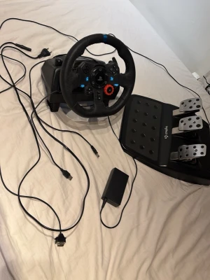 Logitech G29 ratt och pedaler - Säljer ett Logitech G29 racing ratt och pedalset för gaming. Ratten är svart med blå och röda detaljer, har flera knappar och en robust design i plast och metall. Pedalerna är i metall och svart plast, med tre pedaler för realistisk racingkänsla. Perfekt för bilspel!