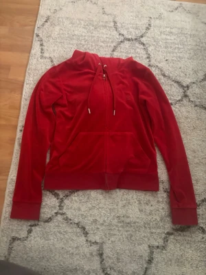 Juicy couture tröja - Snygg röd juicy couture tröja med dragkedja framtill och fickor. Tröjan har huva med dragsko och breda muddar vid ärmslut och nederkant. Perfekt för en chill och avslappnad stil.
