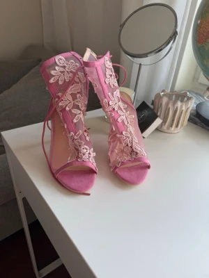 Rosa spetsiga pumps med snörning - Säljer ett par rosa pumps med öppen tå, höga klackar och snörning runt ankeln. Dekorerade med blommig spets i rosa. 10 cm höga