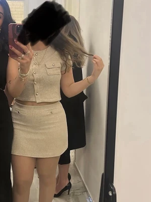 Beige ribbat set med pärldetaljer - Snyggt beige set med ribbad struktur, består av en ärmlös crop top med knapp- och pärldetaljer samt en matchande minikjol. Setet har en stilren och modern look, perfekt för dig som gillar trendiga outfits. Materialet ser ut att vara mjukt och stretchigt. Helt nytt, använt en gång. Köptes för 500-600kr. Passar både S och M 