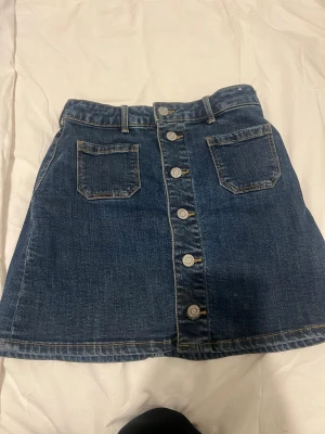 Blå jeanskjol med knappar framtill - Snygg blå jeanskjol i kort modell med knappar hela vägen framtill och två fickor fram. Kjolen har en klassisk denimlook och passar perfekt till vardagsoutfits. Tidlös och enkel att matcha med olika toppar.