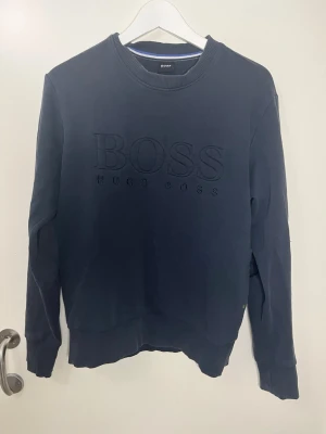 Mörkblå sweatshirt från Hugo Boss - Mörkblå sweatshirt från Hugo Boss med stor BOSS-logga broderad på bröstet. Tröjan har rund halsringning, ribbade muddar och är tillverkad i mjukt bomullsmaterial. Perfekt för en avslappnad och stilren look.