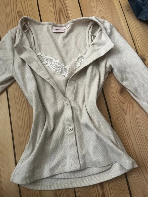 Beige ribbad blus med spets från Gina Tricot - Supersnygg beige ribbad blus från Gina Tricot med knappar framtill och långa ärmar. Blusen har en inbyggd topp med vit spetsdetalj vid urringningen som ger en extra touch. Perfekt att styla med jeans eller kjol för en trendig look.
