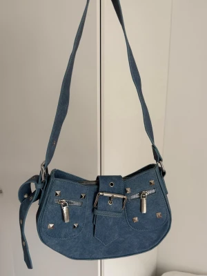 Blå denim axelväska med nitar - Snygg blå axelväska i denim med silverfärgade nitar och detaljer. Väskan har två framfickor med dragkedja, dekorativt bälte och bred axelrem. Perfekt inför våren och sommarfester