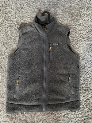 Grå fleeceväst från Patagonia - Mysig grå fleeceväst från Patagonia med hög krage och hel dragkedja framtill. Västen har tre fickor med dragkedja och klassisk logga på bröstet. Perfekt för lager-på-lager och outdoor vibes. Tveka inte på att höra av dig vid frågor! 