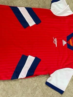 Arsenal Adidas röd matchtröja - Snygg röd Arsenal matchtröja från Adidas med vita ärmar och blå detaljer. Tröjan har klubbens logga på bröstet och klassiska Adidas-ränder i blått och vitt på sidorna. Tillverkad i lätt och ventilerande material, perfekt för träning eller match.