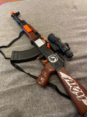 Leksaksvapen AKM cosplay brun/svart - Säljer ett leksaksvapen inspirerat av AKM, perfekt för maskerad eller cosplay. Geväret är i plast med bruna och svarta detaljer, orange markeringar och vit AKM-text på kolven. Utrustad med kikarsikte och bärrem för extra realism.