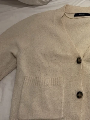 Beige kofta från Vero Moda - Mysig beige kofta från Vero Moda med två stora fickor framtill och knäppning med tre bruna knappar. Koftan har en djup v-ringning och är stickad i ett mjukt material som passar perfekt för lager-på-lager. Enkel och stilren design.