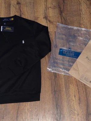 Svart tröja från Ralph Lauren - Snygg svart stickad tröja från Polo Ralph Lauren med klassisk rund halsringning och broderad vit logga på bröstet. Tröjan har ribbade muddar vid ärmslut och nederkant samt diskreta sömdetaljer framtill. Perfekt för en stilren och avslappnad look. Priset går att diskuteras vid snabb affär