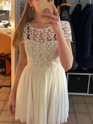 Vit spetsklänning - Söt vit kortklänning med överdel i blommig spets och rund halsringning. Har nån lös tråd på några ställen vid överdelen men annars i fint skick. Kjolen har flera lager. Perfekt för sommarens alla tillfällen och studentfirande. Passar storlek S