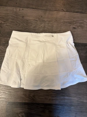 Vit sportkjol från Lululemon - Snygg vit sportkjol från Lululemon med plisserade detaljer och bred midjeresår. Kjolen har en liten ficka bak och inneshorts med fickor på sidorna. Passar bra till tennis, golf eller liknande. Storlek 6 men skulle säga S. Den har en liten nopprig fläck på midja bandet men som inte syns 