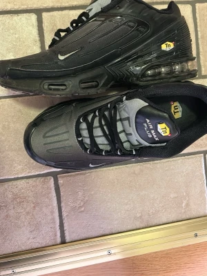 Nike Air Max Plus Tn Svart/Grå - Nike Air Max Plus Tn sneakers i svart och grått med tydliga Tn-detaljer och synlig Air Max-dämpning i sulan. Skorna har en sportig design med mesh och syntetmaterial, samt små vita Swoosh-loggor på sidan. Perfekta för dig som gillar streetwear.