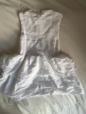 Vit axelbandslös kort klänning - Säljer min vita klänning men volanger på kjolen. Klänningen är i storlek S med hade snarare sagt att den är i xs klänningen är för kort för mig och därav säljer jag den. Den är ifrån Gina och använd en gång på en skolavslutning