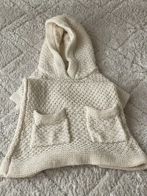 Stickad cremevit poncho med huva - Säljer en stickad cremevit poncho med huva och två stora fickor framtill. Ponchon har ett grovt stickat mönster och ribbstickade kanter. Perfekt för lager på lager och chill stil.