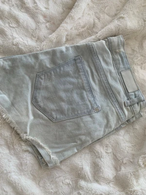 Ljusblå denimshorts med fransig kant - Säljer ett par ljusblå denimshorts med fransig rå kant och klassisk femficksdesign. Shortsen har hög midja och bälteshällor, perfekta för sommardagar. Stilen är loose fit och materialet är bomull.