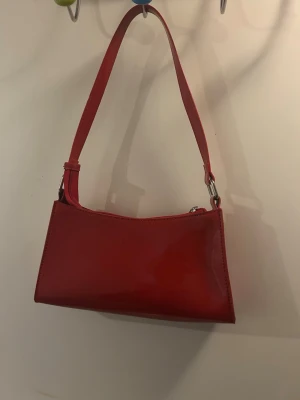Röd axelremsväska i skinnimitation - Trendig röd handväska med axelrem och stilren, rektangulär form. Väskan har en slät yta i skinnimitation och silverfärgade metalldetaljer. Perfekt för dig som vill addera en färgklick till din outfit.