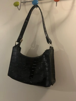 Svart axelväska i krokodilimitation - Snygg svart handväska med axelrem i trendig krokodilimitation. Väskan har en fyrkantig form och stängs med dragkedja. Perfekt accessoar för att lyfta din outfit med en cool och modern touch.