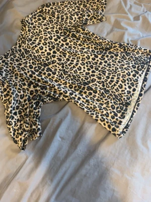 Leopardmönstrad topp med kort ärm - Säljer en snygg topp med leopardmönster i svart och beige. Toppen har rund halsringning och korta ärmar. Perfekt för dig som vill sticka ut lite extra! Materialet känns mjukt och stretchigt, troligtvis polyester.