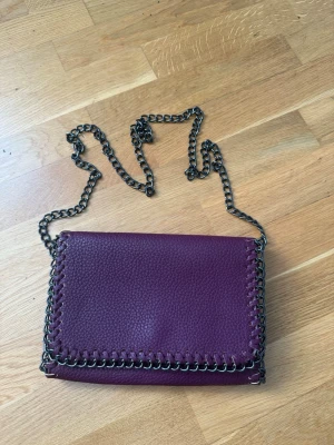 Vinröd axelväska med kedjerem - Snygg vinröd axelväska i konstläder med grov kedjerem och dekorativ kedjedetalj runt kanterna. Väskan har en rektangulär form och lockstängning. Perfekt för dig som vill ha en trendig accessoar med lite edge.