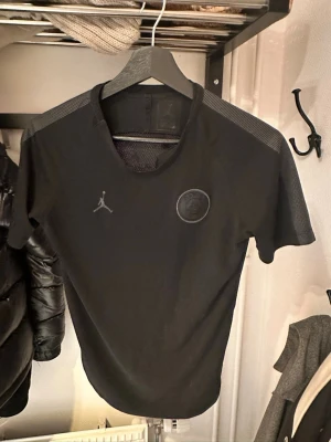 Svart Jordan x PSG t-shirt - Snygg svart t-shirt från Jordan x PSG med diskret logga på bröstet och mesh-detaljer på axlarna. Perfekt för dig som gillar sportig stil och vill ha något bekvämt och stilrent. Klassisk passform och rund hals. Passar XS