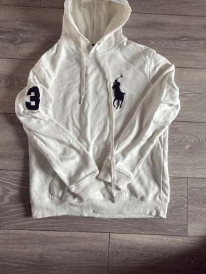 Vit hoodie från Polo Ralph Lauren - Snygg vit hoodie från Polo Ralph Lauren med stor marinblå logga på bröstet och siffran 3 på ärmen. Klassisk känguruficka framtill, ribbade muddar och dragsko i huvan. Perfekt för en clean och sportig look. Inga defekter alls, priset är ej hugget i sten, skriv vid minsta fundering.